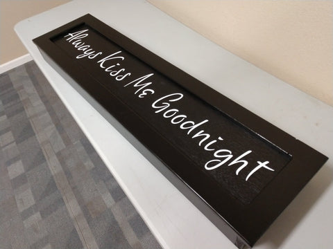 Black wooden wall art with white lettering saying "Always Kiss Me Goodnight" laid on table.