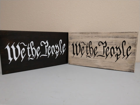 A black, wooden mini decor sign with "We The People" in white script and a white, distressed wooden mini decor sign with black "We The People" script.