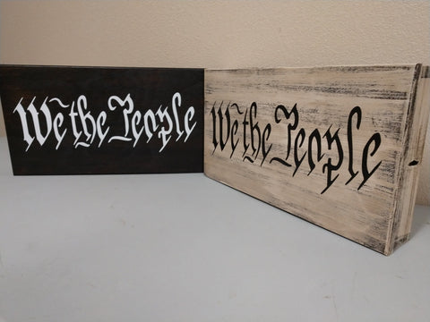 Two "We The People" wall decor signs leaning against a wall.