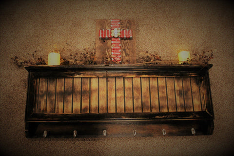 A light brown wooden gun concealment coat rack with two candles and a wooden plaque with a cross made from shotgun shells sitting on top.