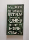 Mini distressed wood wall art with the lettering "All our visitors bring happiness. Some by coming. Others by going." hanging on wall.