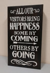 Black, wooden, mini wall art with white lettering saying "All our visitors bring happiness. Some by coming. Others by going." leaning against white wall.