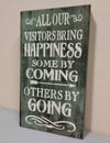 Olive green, distressed mini wooden gun concealment wall art with saying "All our visitors bring happiness. Some by coming. Others by going." leaning against wall.