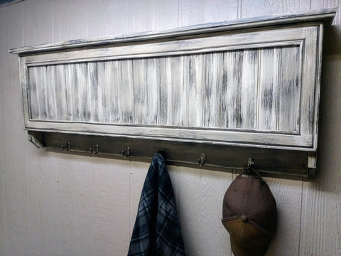 A distressed whitew wooden gun concealment coat rack with a flannel and ball cap hanging on two of its hooks. 
