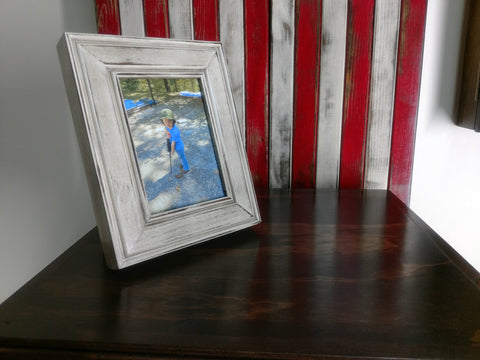 A distressed white 5x7 gun concealment picture frame sitting on the corner of a dark brown wooden table. 