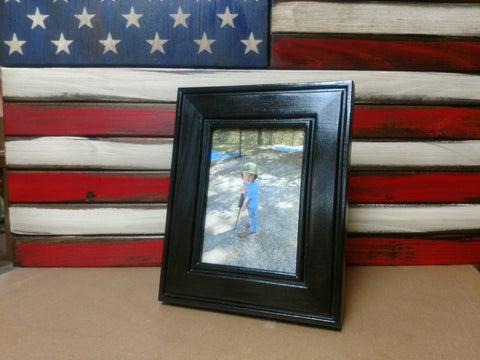 A black 5x7 gun concealment picture frame sitting in front of a red, white and blue concealment flag.