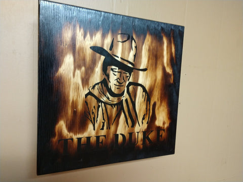 Black and white outline of John Wayne on distressed wooden sign hanging on wall.