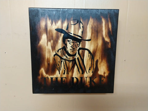 John Wayne outline on distressed wooden sign hanging on wall.