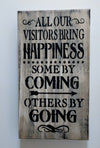 Grey, distressed wooden gun concealment wall art with quote "All our visitors bring happiness. Some by coming. Others by going." hanging on wall.