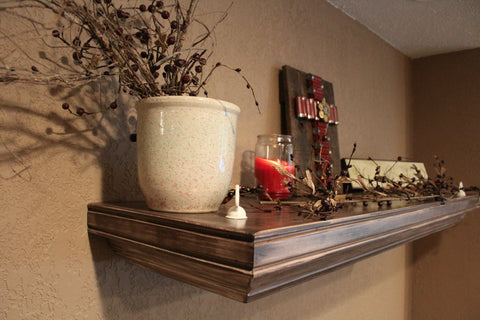 An angled view of a light brown wooden gun concealment shelf with with a candle, ceramic pot and a wooden plaque with a cross made from shotgun shells sitting on top.