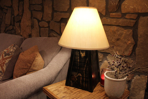 A distressed black wooden gun concealment lamp with a white lamp shade, sitting on a side table next to a grey couch with its light on. 