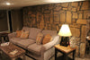 A basement with a stone wall behind a grey cloth couch sitting next to a side table with a wooden gun concealment lamp sitting on it. 